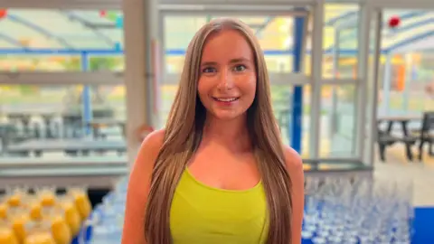 Emily Doughty / BBC Niamh wears a yellow top and is smiling. She is standing in front of a table with glasses and orange juice on