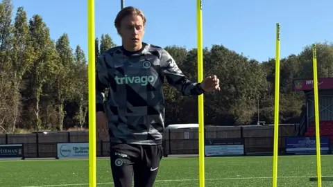 Handout George Edwards wears a grey and black patterned jersey and black Nike joggers. He is dribbling a football on an outdoor pitch. 
