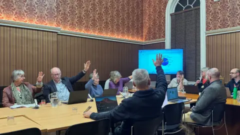 Seth Dellow A group of councillors in a town hall room sitting round a table in front of a screen with a power point on it. Some councillors have their hands raised