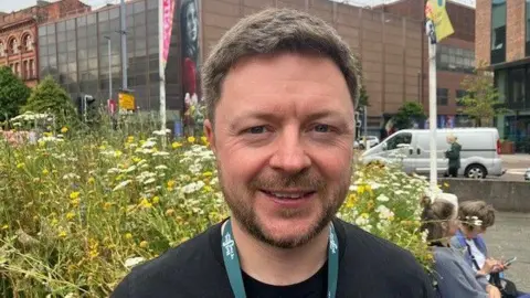 A man with short brown hair and a beard is smiling, with wildflowers behind him as he stands in a city centre park.