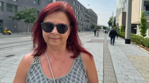 A woman with red hair and sunglasses looks at the camera