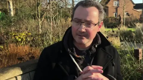 a man sits on a wooden bench in a garden during a BBC interview. He has short grey hair and is wearing glasses and a black winter coat. His hands are clasped together and he is looking downwards, as if thinking about something serious.