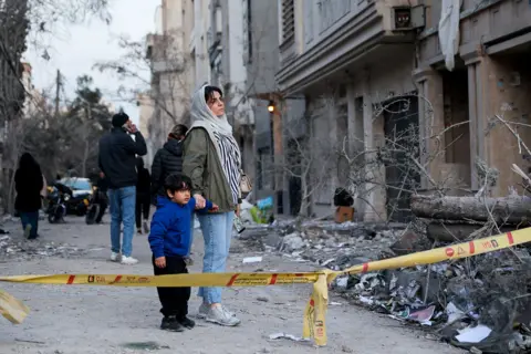 WANA via Reuters A woman with her child stands behind police tape next to rubble and debris on a street and looks at the aftermath of an Israeli and the U.S. strike on a police station, 2 March 2026.