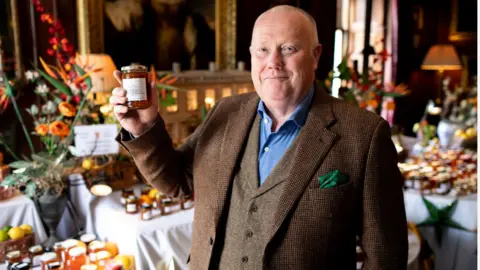 Hermione McCosh An older man in a very smart brown tweed three-piece suit holding up a jar of marmalade and smiling broadly to the camera. He is in a grand room, like in a manor house, and there is a table behind with dozens of marmalade jars laid on it and two elegant lamps casting a yellow glow over them