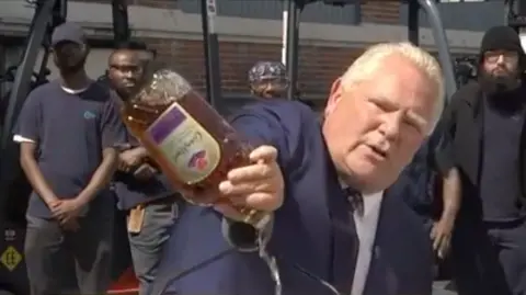 Premier Doug Ford pouring down a bottle of Crown Royal whiskey in front of a podium. He is wearing a suit and tie, and has short light blonde hair.