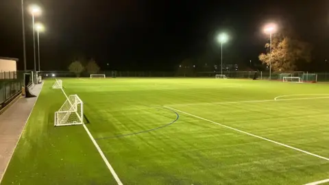 Matt Cutler View of brand-new AstroTurf on a football pitch. It is dark and the floodlights are on.