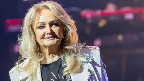 Bonnie Tyler on stage, she has long blonde hair, and is wearing a black T-shirt and a white biker jacket