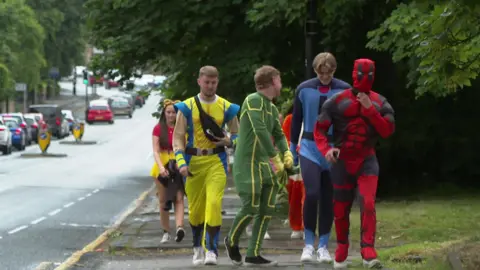 A group of young adults walk down a street in superhero fancy dress. 