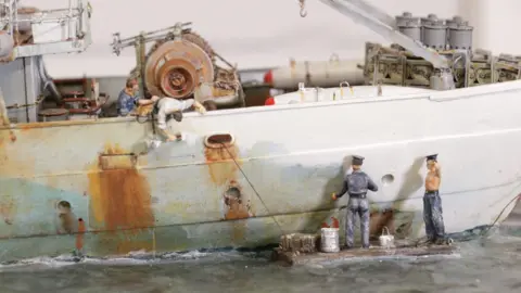 Elliott's Auctioneers Models of sailors paint the side of slightly rusty ship which is still at sea