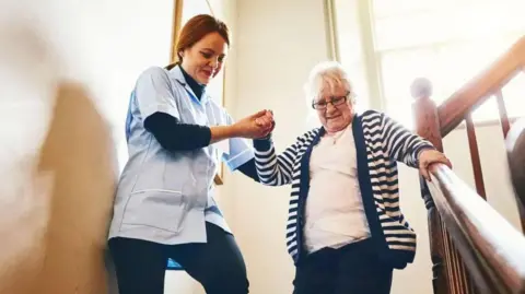 Getty Images A care assistant helping an elderly woman to walk down a flight of stairs. 