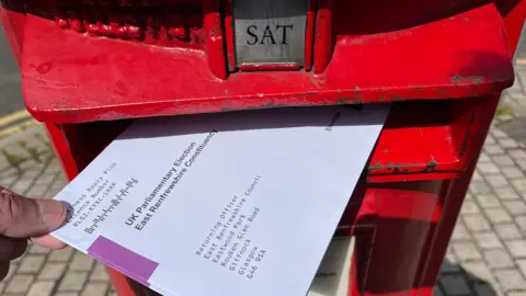 BBC A red post box with an envelope addressed to East Renfrewshire Council's Returning Officer, containing a postal ballot 