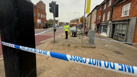 Jamie Niblock/BBC A police officer stands in front of an area cordoned off with blue and white police tape. Everything sits on a sidewalk next to a road with shops. It is a gray and cloudy day.