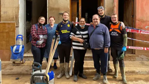 Zoe Wilkes A group of volunteers wearing wellies and gloves, standing in front of a building with three open doors in Valencia. They are posing with their arms around each other and smiling at the camera. They are there to help with the clean up efforts. In front of them there is a bucket full of brooms and cleaning supplies.