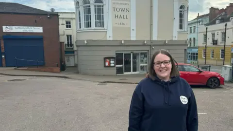 A woman with glasses stood in front of a pale cream and grey building with Lutterworth Town Hall written on the side 