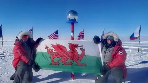 Georgina Gilbert and Rebecca Openshaw-Rowe posing with a Welsh flag at the ceremonial South Pole.