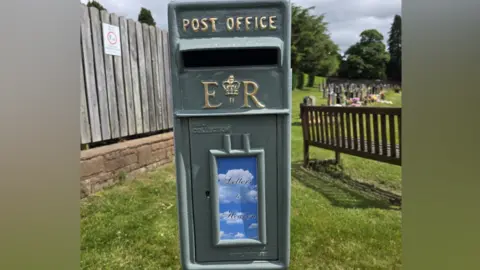 A dark green post box sits in a cemetery. It has a sign on the front with 'letters to heaven' against a blue sky and clouds. 