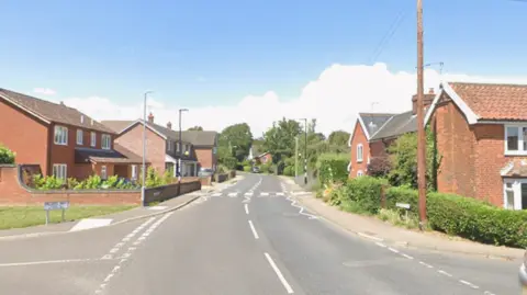 A residential street with a broken white line down the middle of the road. It is a sunny day with a blue sky. Red brick houses line either side of the street. A pedestrian crossing is marked on the road and junctions go off to the left and right in the foreground.  
