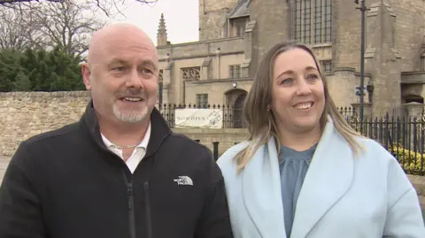 Simon and Tanya Brown standing side by side outside a large sandstone building with decorated windows and spires. Mr Brown is bold and has a grey goatee. He is wearing a black zip-up fleece over a whites shirt and he is wearing a silver chain around his neck. Mrs Brown has shoulder-length light brown hair and is wearing a baby blue coat over a grey-blue blouse. They are speaking to a reporter who is interviewing them out of shot.