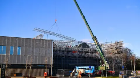 Cumberland Council A crane is lifting a steel girder off the roof of the Sands Centre while a man in high vis jacket watches. It's a large brown building covered in scaffolding. 
