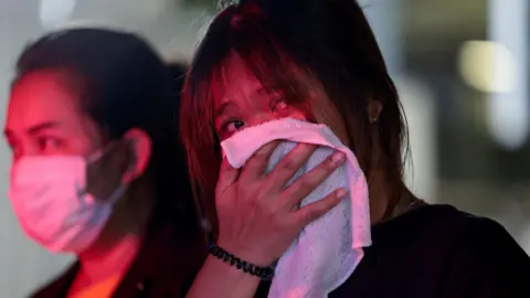 Two women stand, their faces lit by emergency services lights, one wearing a face mask and the other holding a white cloth over her mouth and nose. 