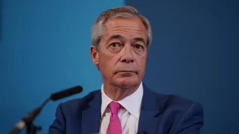 Nigel Farage is sat down in front of a microphone and a blue backdrop. He has short grey hair and is wearing a dark blue suit, white shirt and a pink tie.