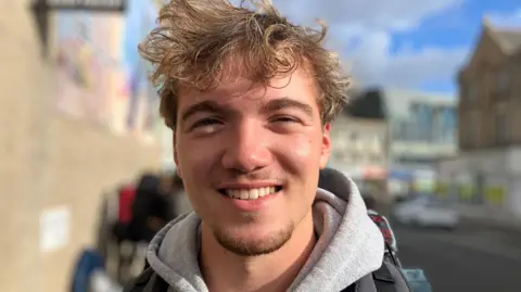Will has brown hair and is wearing as grey hoodie and is smiling at the camera.