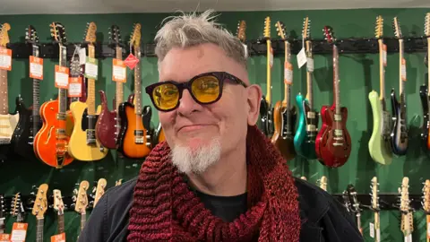 ELLEN KNIGHT/BBC Alan photographed looking into the camera and smiling slightly. He's wearing a black shirt, and has a red knitted scarf on. He's wearing black-framed glasses that have yellow-tinted lenses. He has short grey hair and a beard. He's stood in front of a dark green wall, which has two rows of guitars - in colours ranging from bright orange to light green - hanging up behind him. 
