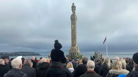 The back of people's heads as they look at the Douglas war memorial, which is a tall war stone memorial with a statue of a solider on the top. The sea and Onchan Head can been seen in the background.