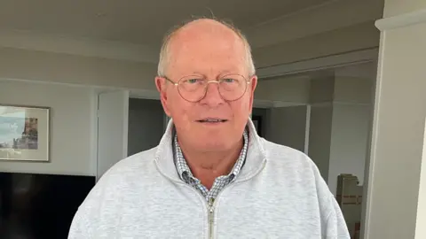 A man in a grey fleece stands in his living room. He has short white hair and glasses. He is faintly smiling. 