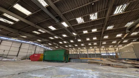 Suffolk County Council The inside of a large, abandoned warehouse-style building. There is a large green container, a red and yellow skip, and large bits of wood on the floor.