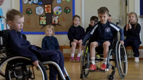 Jamie Niblock/BBC Two boys wearing navy school uniform riding wheelchairs in a school hall. They are cheered on by other pupils sat on a bench behind them.