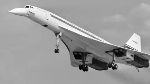 A black and white image of Concorde in flight. It has a pointed nose and thin body with smooth, curved wings.