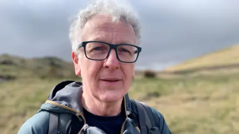 A man with grey hair and dark rimmed glasses is looking at the camera. He is standing in front of a rugged area. He is wearing a blue coloured waterproof jacket with and has a rucksack on his back.