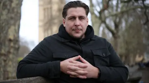 Reuters Joe Bennett, who has combed back brown hair and wears a black buttoned-up jacket, is seen folding his fingers and has a neutral expression in a profile shot
