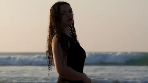 Toby Piper A young-woman with long wavy brown hair, in dark lighting with red lipstick on. She has dark eyes and a dark dress on, and is stood on a beach in front of a breaking wave.