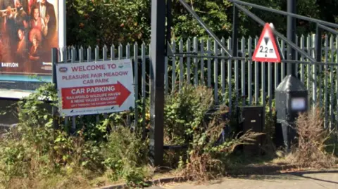 Google Maps A Google Street View image showing a sign for Pleasure Fair Meadow car park attached to green fencing which is surrounded by overgrown weeds and plantation