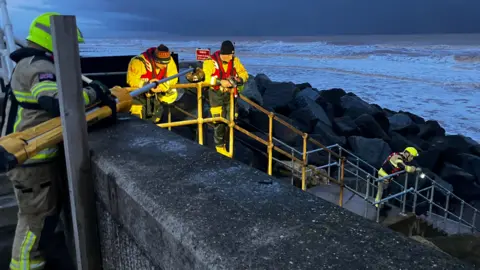 BBC/Jessica Lane Four members of the coastguard are looking over the steps onto the rocks on the beach. One is leaning forward looking with a torch.