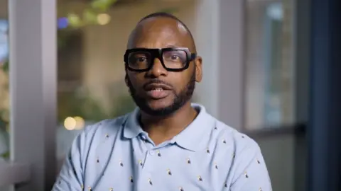 Christopher Joell-Deshields looks at the camera seriously, he is wearing a baby blue polo shirt with tiny puffins on it. His glasses are thick-rimmed black glasses and his hair is cropped short.