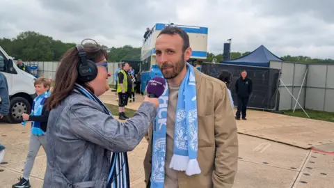 Liam Kelly is interviewed wearing a beige jacket and wearing a Coventry City light blue scarf.