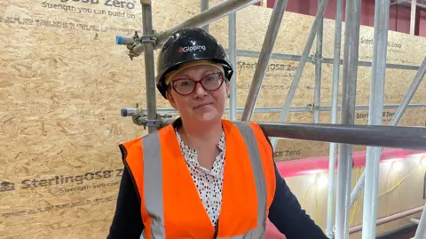 Shaun Whitmore/BBC A photo of museum manager Alison Hall. She is smiling to camera while wearing a black safety helmet and orange high-vis jacket. Around her is scaffolding along with plywood construction boards protecting the museum's walls.