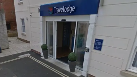 The doorway of Ryde Travelodge is flanked by ornamental shrubs in pots with a blue company sign above it.