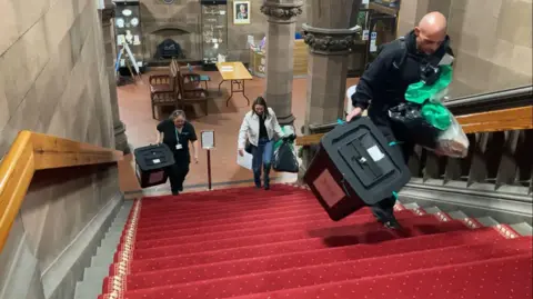 There is a staircase and the picture is taken from the top of it. The carpet on the stairs is red and there are three people walking up the stairs. They are carrying ballot boxes and bags. One is a bald man, the other two are women. One is wearing a white coat and the other a black polo shirt. At the bottom of the stairs there are some wooden seats and grey stone columns.