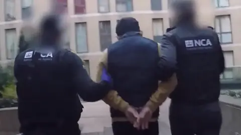 NCA Le is led away from his flat by two NCA officers in uniform. He has his hands cuffed behind his back and his back to the camera as do the officers.