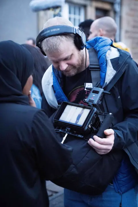 Compass Collective A man with a beard and wearing headphones is checking the viewfinder of a camera. He is with a young woman wearing a hijab. We can only see the back of her head.