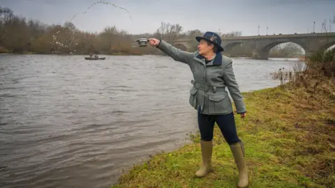 Sarah-Jane Laing, chief executive of Scottish Land & Estates, is standing on a bank of the Tweed. She is wearing a black fedora-type hat, grey jacket, black leggings and green boots. She is throwing whisky into the river from a quaich.