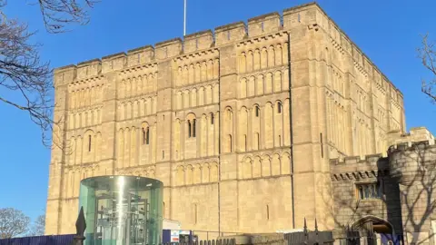 Shaun Whitmore/BBC The exterior of Norwich Castle. It is a cube-shaped building made of pale brown stone. A path with railings leads to a smaller, attached part of the castle next to the cube. There is a see-through cylinder structure in front of the castle, containing metal machinery.