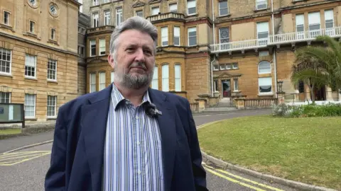 Mark has grey hair and a beard. He is wearing a blue and green striped shirt with a dark blue jacket
