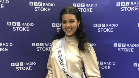 A woman wearing a pale-coloured top and a white sash is standing against a purple BBC Radio Stoke backdrop. She has curly black hair and is smiling for the photograph.