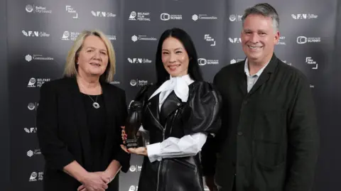 Press Eye Lucy Liu poses for a photo while standing between Belfast Film Festival director Michele Devlin and Richard Williams from Northern Ireland Screen. Ms Devlin has shoulder-length blonde hair and is wearing a black suit and top. Mr Williams has short, greying hair and is wearing a dark jacket over a pale-coloured open-neck shirt. 