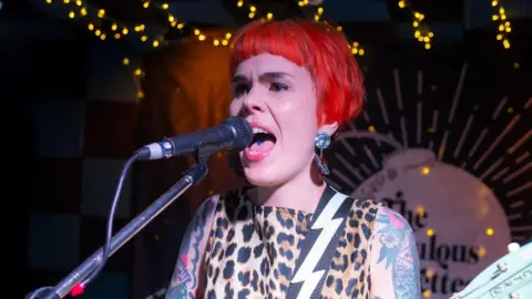 A woman with short red hair sings into a microphone on stage. She is wearing a leopard print dress with a lightning-print guitar strap. She has many tattoos. 